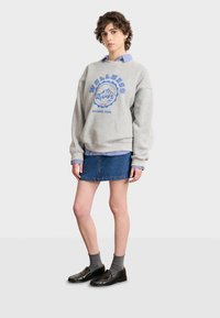Sudadera gris de gran tamaño con texto y gráfico en azul, superpuesta sobre una camisa de rayas en azul claro. Combinada con una falda de mezclilla y zapatos negros.