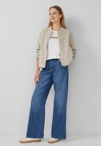 Veste bomber gris clair ; t-shirt blanc avec accent métallisé ; jeans larges bleus ; baskets beiges avec accents blancs ; tenue décontractée.