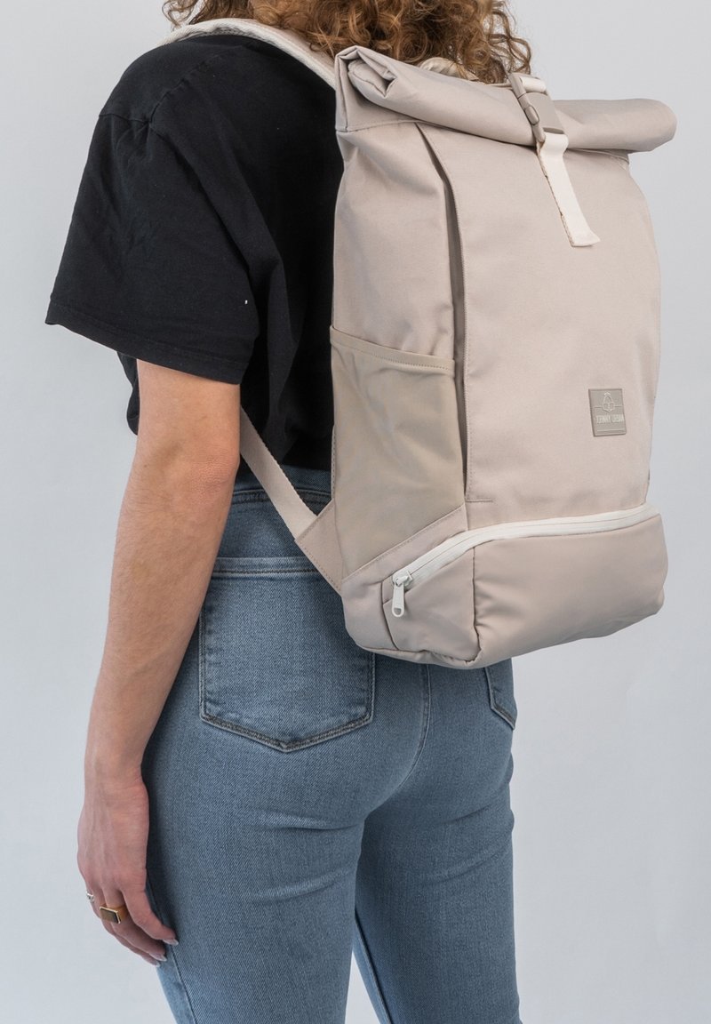 Personne portant un t-shirt noir, un jean bleu et un sac à dos beige à fermeture roulante avec poche zippée à l’avant, vue de côté.