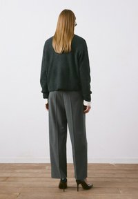 Mujer con cabello largo, gafas, suéter negro, pantalones grises de pierna ancha y tacones altos negros, de pie sobre un suelo de madera frente a una pared blanca.