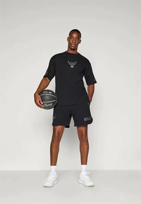 Juodas trumpas rankovėmis marškinėlis su buliuko logotipu, derinamas su juodomis šortais, ant kurių užrašyta "BULLS". Modelis laiko krepšinį, vilkintis baltus sportinius batelius.