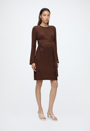 Femme portant une robe marron côtelée à manches longues, longueur genou, avec une ceinture à la taille et des sandales blanches à talons ouverts, debout sur fond blanc.
