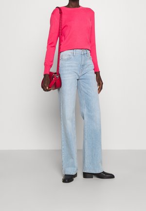 Felroze trui, lichtblauwe wijde jeans en zwarte enkellaarsjes. De persoon draagt een kleine rode crossbodytas.