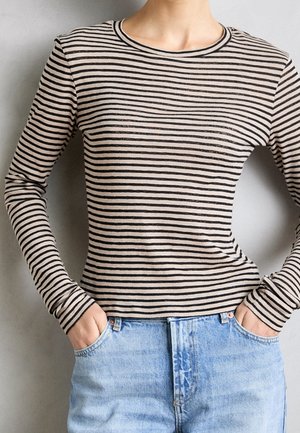 Persoon draagt een beige en zwart gestreept shirt met lange mouwen, ingestopt in lichtblauwe spijkerbroek, met handen in de zakken.