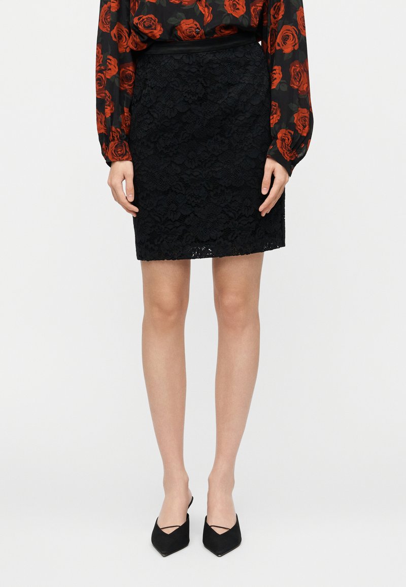 Jupe crayon en dentelle noire, avec tissu texturé en couches, coupe ajustée et ourlet festonné. S'associe avec un chemisier noir et des talons.