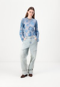 Prosojno modra bluza z dolgimi rokavi in cvetličnim vzorcem, v kombinaciji s svetlimi širokimi džinsom. Model nosi temne gležnjarje na peto.