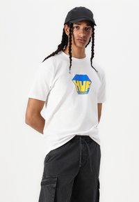 Hvid bomulds T-shirt med et flerfarvet "HUF" logo. Bæres med sorte cargo bukser. Modellen har en sort kasket på og har flettet hår.