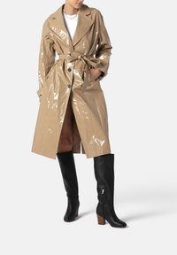 Trench coat beige lucido con vita legata, ampi revers e dettagli con bottoni, abbinato a stivali alti fino al ginocchio in pelle nera con tacco a blocco.