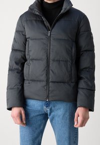 Giacca nera imbottita con texture a scacchi, zip frontale e cappuccio. Abbinata a jeans azzurri per un contrasto, che mette in risalto il design della giacca.