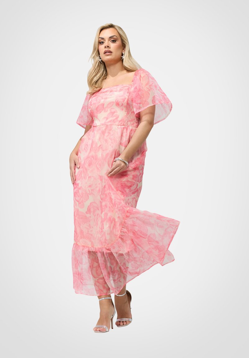 Femme portant une robe midi fluide rose à fleurs avec des manches volantées et des sandales argentées à talons hauts, debout devant un fond uni.