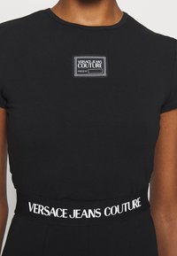 Svart kroppsnära t-shirt med rund halsringning, prydd med en vit logotyp-patch på bröstet och en kontrasterande vit märkesresår.