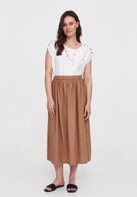 Biała bluzka z guzikami na ramionach, brązowa plisowana spódnica midi i czarne plecione sandały. Prosty design, luźny krój i gładka tekstura.