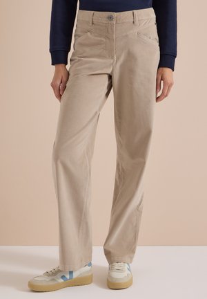 Pantalon beige droit porté avec des baskets blanches avec des touches bleues et des semelles en gomme, associé à un haut à manches longues foncé.