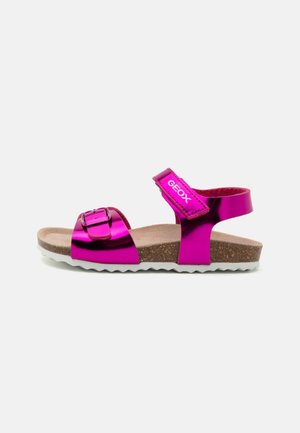 Sandale pour enfants en métal rose vif avec des sangles réglables à la cheville et aux orteils, semelle en liège et semelle texturée blanche.