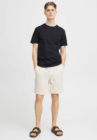 Camiseta de algodón negra con mangas cortas y un bolsillo en el pecho, combinada con pantalones cortos color beige claro y sandalias de ante marrón.