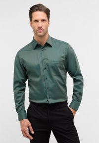 Donkergroene, langmouwige blouse met knopen en een zak, met een textuurafwerking en zwarte knopen, gecombineerd met zwarte broek.