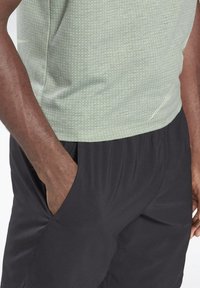 Hombre de piel oscura con camisa ligera de puntos verdes y pantalones negros, con la mano izquierda en el bolsillo, contra un fondo blanco.