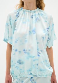 Ljusblå blus i flödande tyg med en rynkad halsringning, korta ärmar och en elastisk nederkant med ett subtilt akvarellmönster.