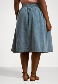 Jeanskjørt med innsnørt midje, A-linjeform, lyseblå farge og en kantlinje som faller under kneet. Brune sandaler synlige.