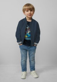 Granatowa kurtka bombowca z białymi paskami, koszulka z grafiką przedstawiającą piłkę nożną, niebieskie dżinsy i białe tenisówki. Gładki materiał i swobodny krój.