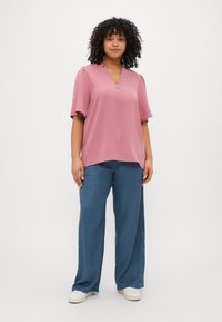 Pink, short-sleeve blouse with a V-neck and relaxed fit. Paired with light blue, wide-leg pants and white sneakers. Simple, textured fabric.