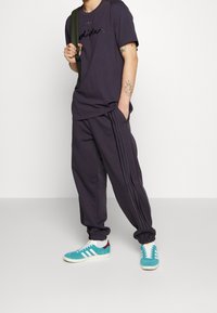 Camiseta gris oscuro con logo negro, pantalones de chándal grises a juego con franjas laterales, y zapatillas en teal con acentos en blanco y marrón.