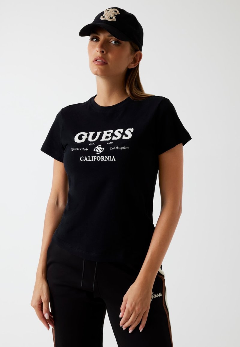Sort bomulds t-shirt med "GUESS" logo i hvid, parret med sorte joggers med sideaccenter og broderet logo. Kapseldetaljer synlige.