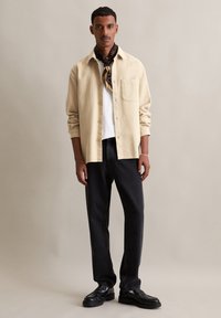 Beige getextureerde button-up shirt over een witte t-shirt; zwarte recht toegepaste jeans; zwarte loafers; gemusterde donkere sjaal geknoopt om de nek.