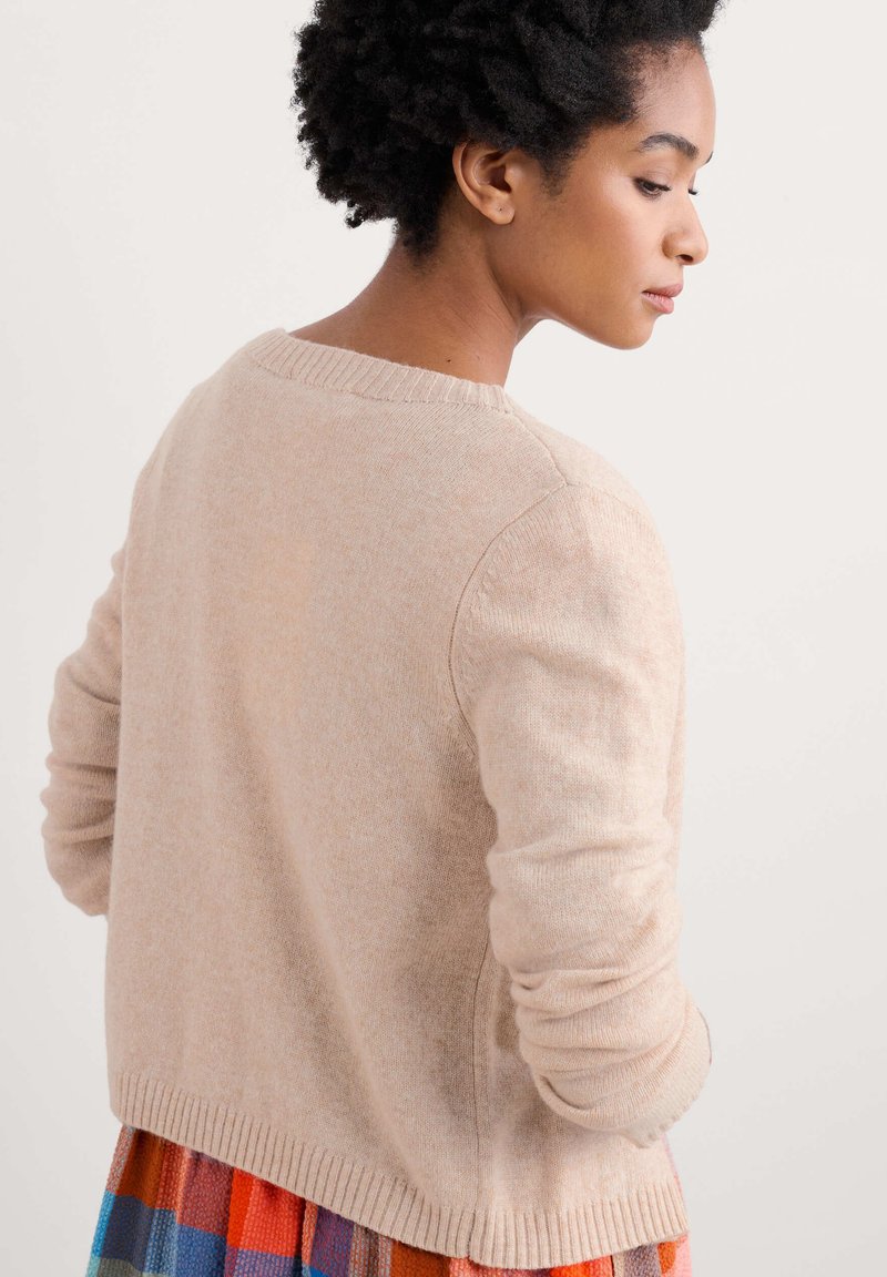 Beiger Strickpullover mit langen Ärmeln, geripptem Halsausschnitt und unterem Saum, der eine weiche Textur und eine lockere Passform aufweist, getragen über einem bunten Rock.