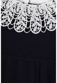 Col blanc en dentelle avec des motifs floraux et de feuilles sur un fond de tissu tricoté foncé texturé.