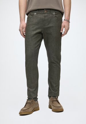 Homme portant un pantalon slim vert olive, des chaussures en suède taupe à lacets et une chemise taupe, debout devant un fond clair uni.