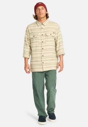 Tan patterned flannel shirt with two chest pockets and button closures, paired with green corduroy pants and navy sneakers.