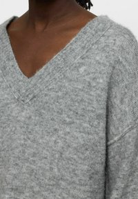 Pull gris tricoté avec un col en V et une coupe décontractée. Texture douce, aspect légèrement duveteux, avec des détails côtelés au niveau du col et des manches.