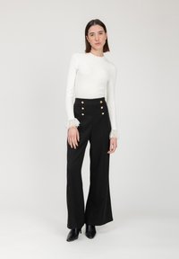 Witte aansluitende lange mouw top met kantaccenten bij de manchetten, gecombineerd met hoge zwarte wijde broek met gouden knoopdetails.
