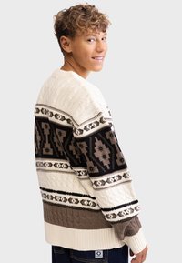 Strikket sweater i creme, brun og sort. Har mønstrede bånd med geometriske designs. Kabelstrik tekstur og ribbede manchetter.