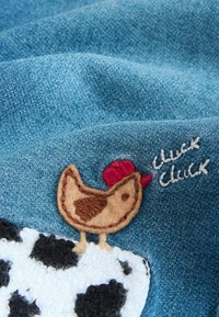 Sinistä farkkukangasta, jossa ruskea huovutettu kana punaisessa hatussa seisoo valkoisella ja mustalla kuvioidulla lehmän läiskällä, jossa teksti "chuck chuck".