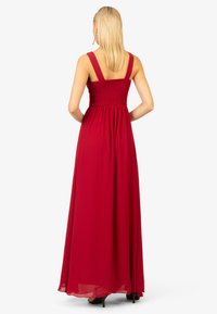Lungo vestito rosso con spalline larghe, busto smockato e gonna fluida. Realizzato in tessuto leggero, presenta una texture liscia e una caduta elegante.