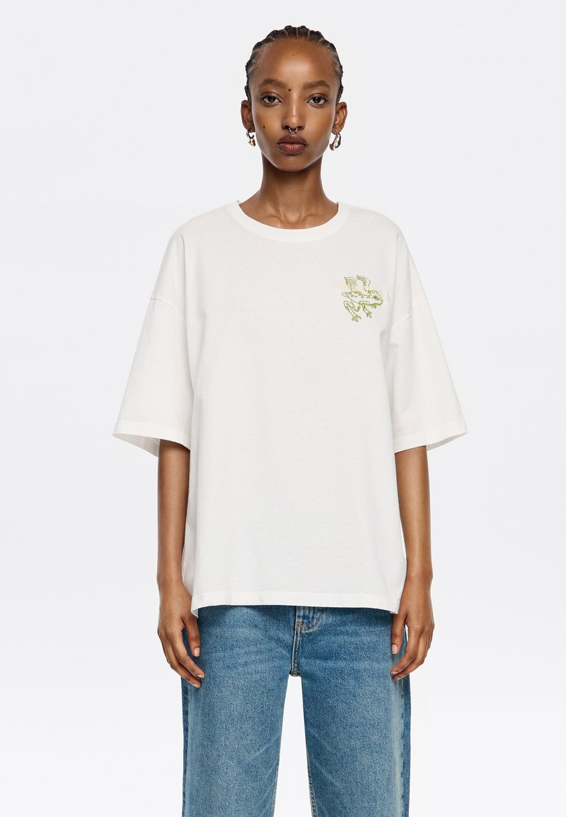 Camiseta de algodón blanca de manga corta con cuello redondo y un estampado gráfico verde en el pecho. Corte holgado, combinada con jeans de mezclilla azul.