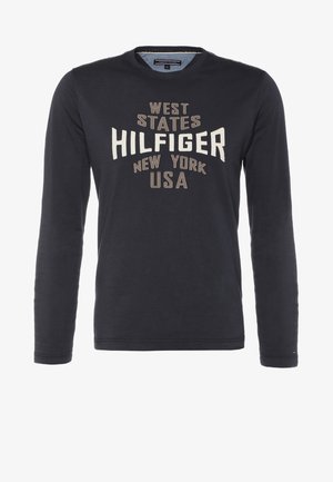 Granatowa koszula z długim rękawem z grafiką "Hilfiger", khaki spodnie i czarne buty z czerwonymi sznurówkami. Torebka na ramię w dłoni, w pozie spacerowej.