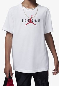 Vit t-shirt med "JORDAN" i svarta bokstäver och en röd Jumpman-logotyp. Modell som bär svartspräckliga byxor och håller en röd väska.