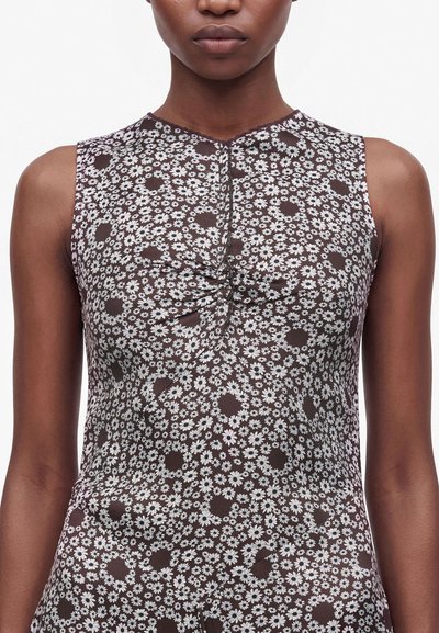 Femme portant un haut marron sans manches avec un motif floral marguerite blanc et un léger froncé au centre devant.
