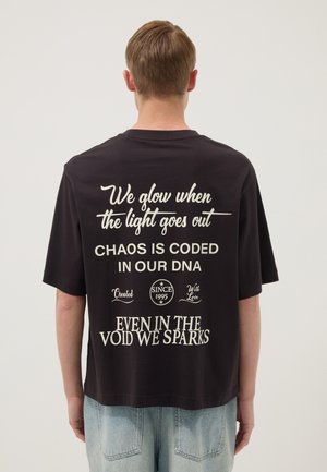 Persona con cabello corto castaño claro lleva una camiseta negra con texto inspirador blanco en la espalda y vaqueros azul claro.