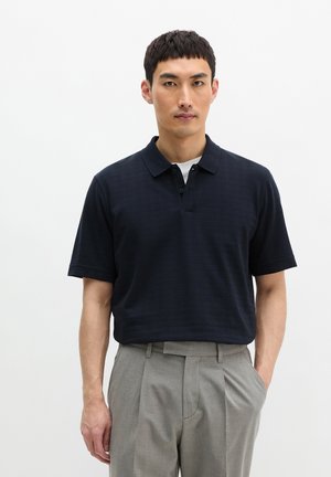 Mann trägt ein marineblaues Poloshirt über einem weißen Unterhemd und graue, plissierte Hose, steht mit einer Hand in der Tasche vor weißem Hintergrund.