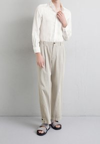 Biała koszula z długim rękawem o subtelnym wzorze, beżowe szerokie spodnie z elastycznym paskiem oraz czarne sandały z szarymi paseczkami.