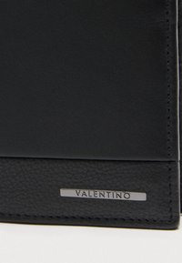 Črni usnjeni predmet z gladkimi in grobozrnastimi teksturami, ki ima majhno pravokotno kovinsko ploščico z vgraviranim napisom "VALENTINO" blizu roba.