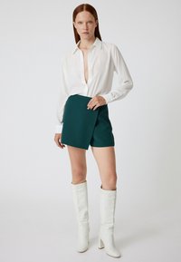 Weißes Langarm-Hemd mit Knopfleiste, grüne Wickelhorts und knietiefel hohe weiße Stiefel mit Absatz. Glatte Texturen und taillierte Passform.
