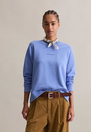 Lichtblauwe sweatshirt met ribbelboorden en ronde halslijn, voorzien van kleine tekstdetails aan de voorkant. Gecombineerd met een paarse corduroy broek en een riem.
