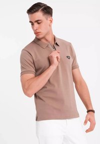 Polo en algodón marrón claro con cuello con cremallera, mangas cortas y un parche de logo negro en el lado izquierdo del pecho.