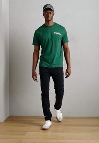 Green cotton T-shirt with a crew neckline, paired with dark jeans and white sneakers featuring gray accents. Model walking against a neutral background.