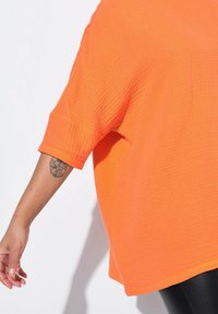 Top arancione con texture, a maniche corte, caratterizzato da una vestibilità comoda. Il tessuto presenta un motivo rigato, arricchito da un tatuaggio visibile sul braccio.